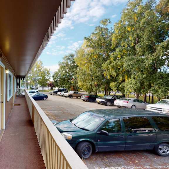 outdoor hallway here at country lane apartments with wooden siding, overlooking a parking lot. several cars are parked next to lush green trees under a partly cloudy sky.