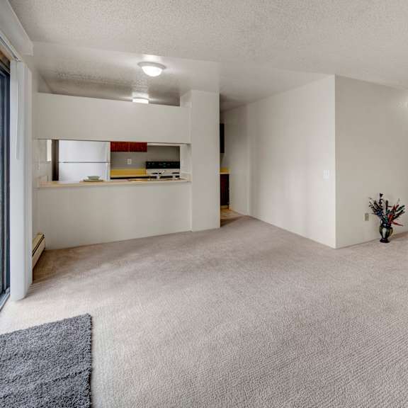 spacious, empty living room here at deer park apartments with light beige carpet, white walls, and a small vase with dried flowers. kitchen with white appliances in the background.