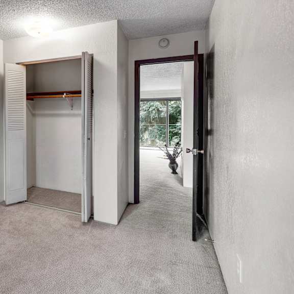 a carpeted bedroom here at deer park apartments featuring an open closet and a doorway leading to a light-filled space with a window and potted plant.