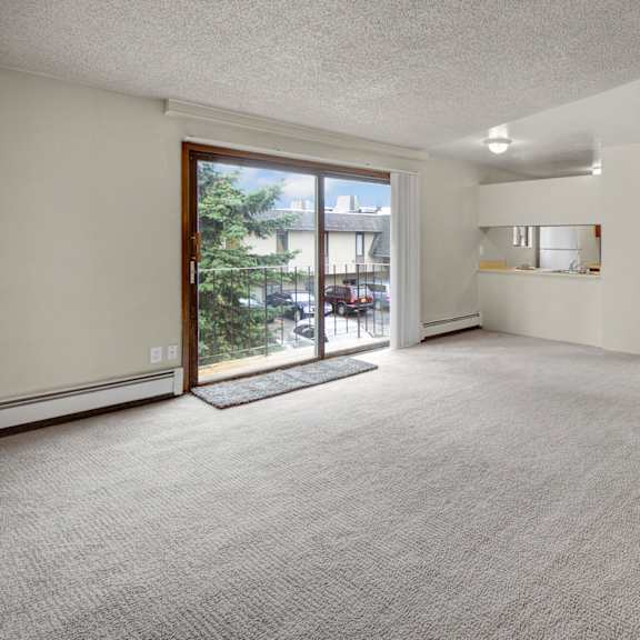 spacious, well-lit living room here at deer park apartments with beige carpet, sliding glass doors leading to balcony, and an adjacent kitchen. a vase with colorful branches sits in the corner.