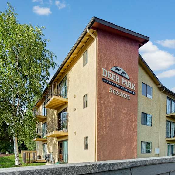 exterior view of deer park apartment homes with balconies, a sign displaying the name and contact number, surrounded by greenery and blue sky.