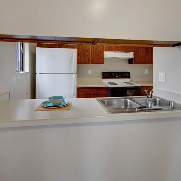 minimalist kitchen here at deer park apartments with a clean white countertop and a double sink, stainless steel faucet, white refrigerator, stove, wood cabinets, and a neatly placed blue dish set.
