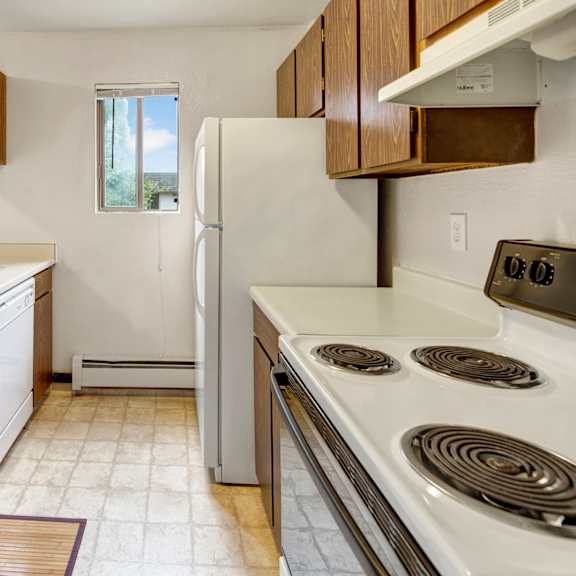 compact kitchen here at deer park apartments with wood cabinets, white appliances, and a small window. includes a stove, double sink, and dishwasher. bright, clean, and orderly.