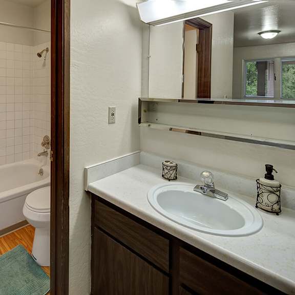 bathroom here at driftwood apartments with a white-tiled bathtub and shower curtain, wooden floor, and vanity sink. soft lighting and a calm, clean atmosphere.