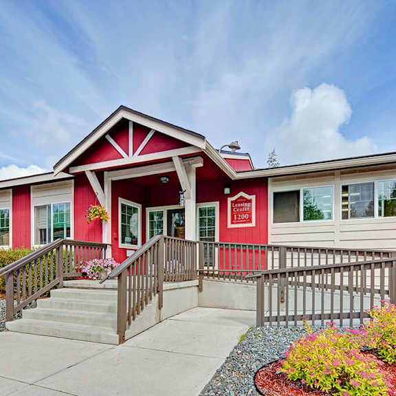 front view of a red and white building here at the glen apartments, identified as a leasing center. the entrance features steps and a ramp, surrounded by colorful landscaping under a partly cloudy sky.