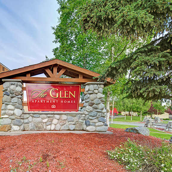 stone sign for "the glen apartment homes" near a tan building, surrounded by lush greenery and pine trees, under a clear, blue sky. calm atmosphere.