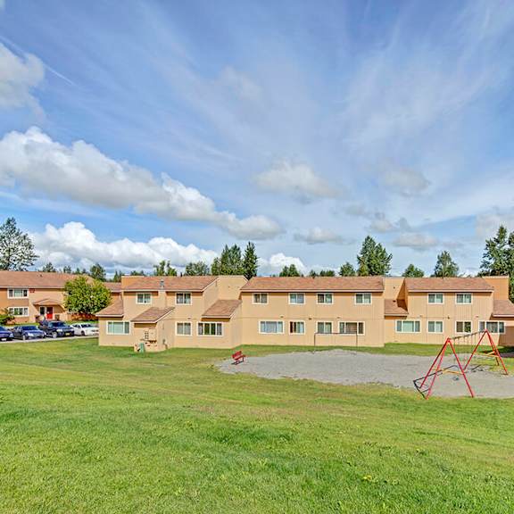 the glen apartment complex under a bright blue sky with clouds. yellow buildings have brown roofs and are surrounded by green grass. a red swing set is visible.