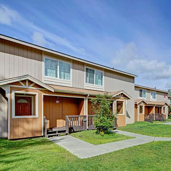 two-story beige townhouses here at the glen apartments with porches, surrounded by green lawns and trees on a sunny day. the atmosphere is peaceful and suburban.