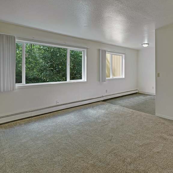 a bright living room here at the glen apartments with beige carpet and a large window showing green trees. white walls, partial view of a kitchen with a fridge. calm, open atmosphere.