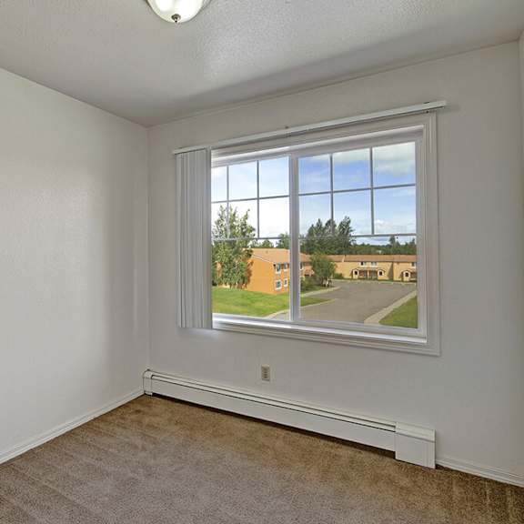 bedroom here at the glen apartments with white walls, carpeted floor, and a large window revealing a view of houses, greenery, and a blue sky. calm and clean atmosphere.