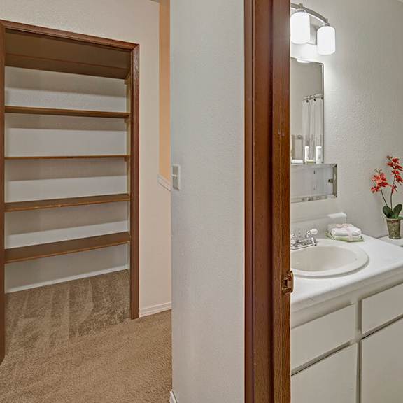 a clean bathroom here at the glen apartments with white fixtures, including a sink, toilet, and bathtub with a patterned curtain. a small red plant decorates the vanity. on the left, an empty closet with wooden shelves is visible, creating a tidy and minimalistic atmosphere.