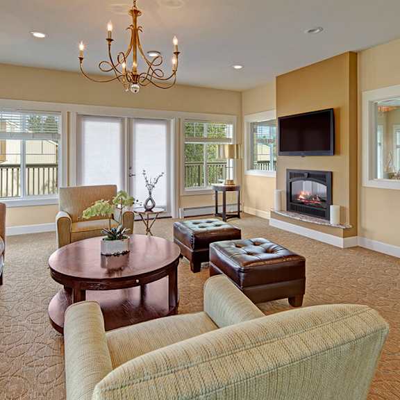 cozy living room here at the glen apartments with a modern design. features beige couches, a round wooden table, a fireplace, and large windows. a chandelier adds elegance.