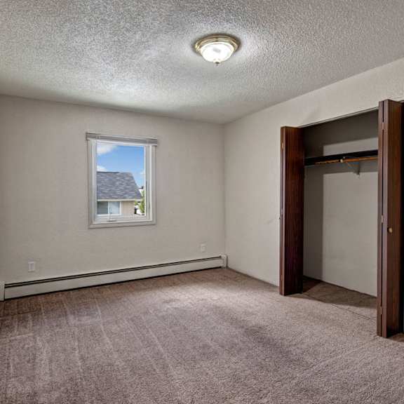 bedroom here at greenbriar apartments with beige walls and brown carpet. a single window shows a roof and sky. open closet with wooden doors on the right. simple and neutral tone.