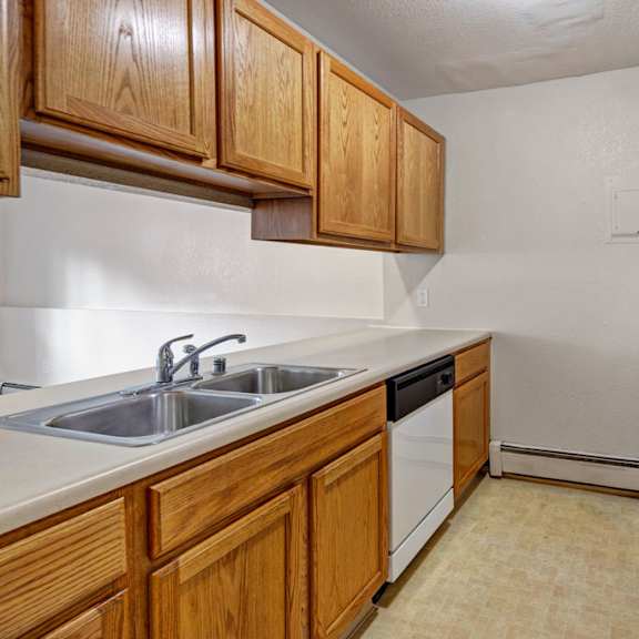 narrow kitchen here at greenbriar apartments with wooden cabinets, beige countertops, and appliances including a stainless-steel sink, dishwasher, white stove, and fridge. cozy and functional.