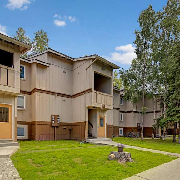 two-story beige apartment buildings here at greenbriar apartments with brown trim amidst a landscaped, grassy area with trees and blue sky, conveying tranquility.