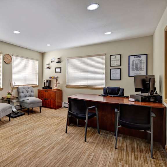 office here at greenbriar apartments with a wooden desk, black chairs, and a computer. two gray armchairs and a potted tree add warmth. certificates and a clock adorn the walls.