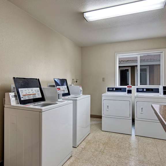 a clean, brightly-lit laundry room here at greenbriar apartments with four white washing machines and dryers lined against cream-colored walls. a window reflects natural light.