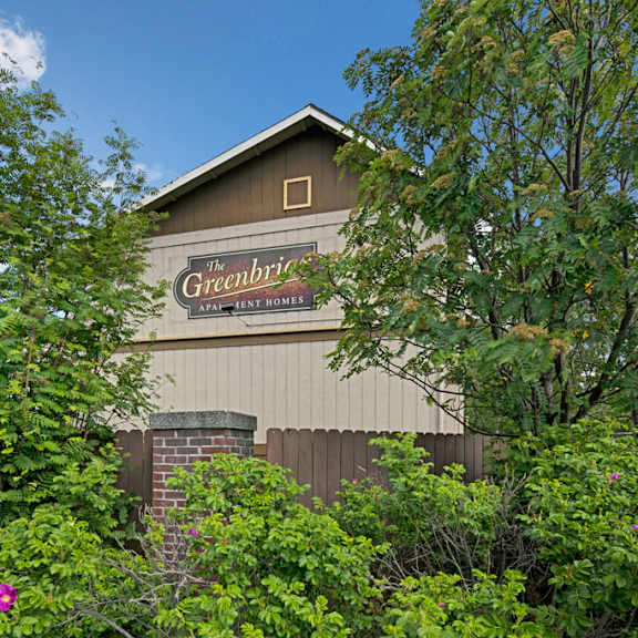 a building sign for "the greenbriar apartment homes" is partially obscured by lush green trees and shrubs, with a clear blue sky in the background.