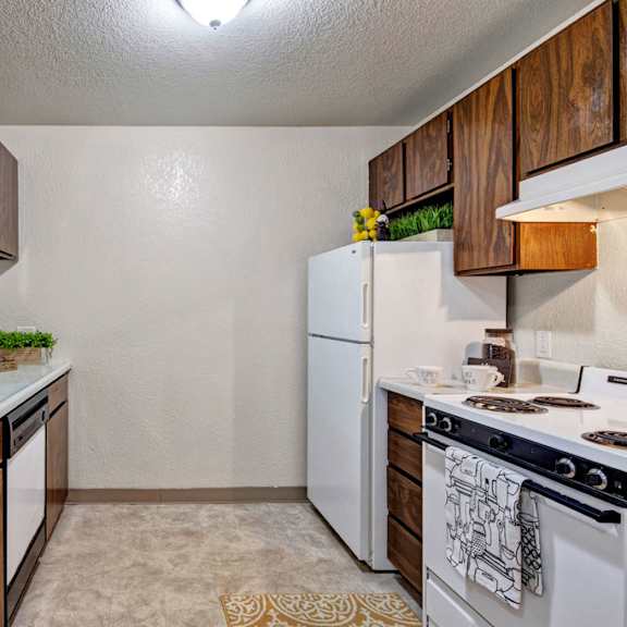 compact kitchen here at greenbriar apartments with dark wood cabinets, white appliances including a fridge and stove, dual sink, and decorative green plants. cozy and functional.