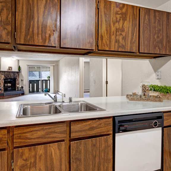 stylish kitchen here at greenbriar apartments with wooden cabinets, double sink, and dishwasher. a pass-through window reveals a cozy living room with a fireplace, creating a warm tone.