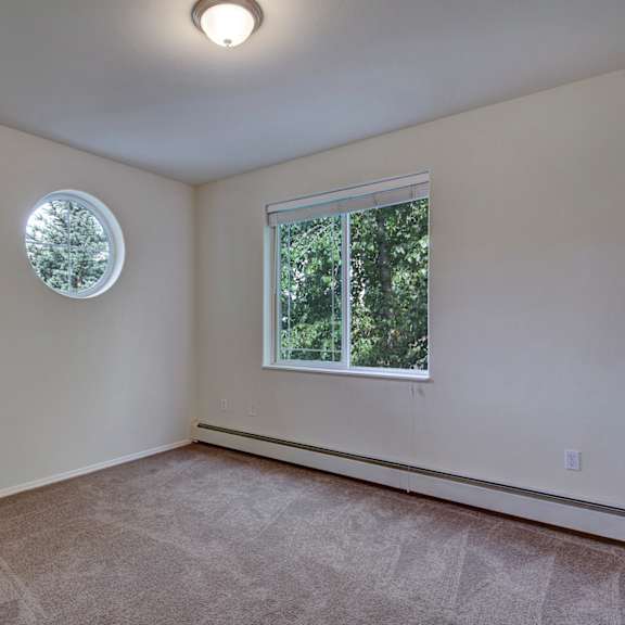 an empty room here at the highlands luxury apartments with beige carpet, white walls, and two windows. a round window shows a tree outside, and a larger window has blinds, offering greenery views.