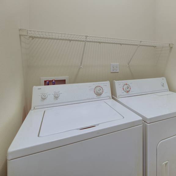 a laundry room her at the highlands luxury apartments with a white washer and dryer side by side. above them is a wire shelf. the walls are light beige, creating a clean, tidy feel.