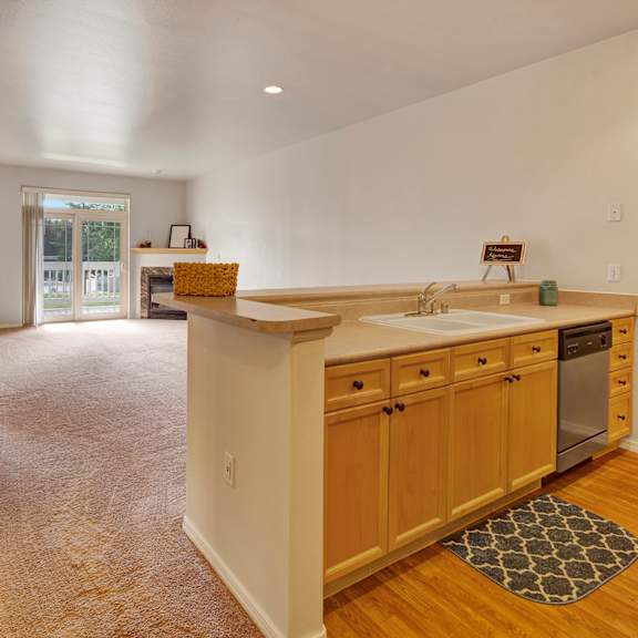 open-plan living space here at the highlands luxury apartments with light wood kitchen cabinets, a sink, and dishwasher. cozy carpeted area leads to a sunlit balcony, creating an inviting ambiance.
