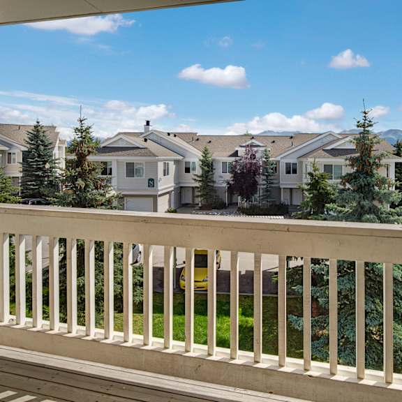 view from a balcony here at the highlands luxury apartments overlooking a row of townhouses with a bright blue sky and scattered clouds. tall pine trees and parked cars line the street below.
