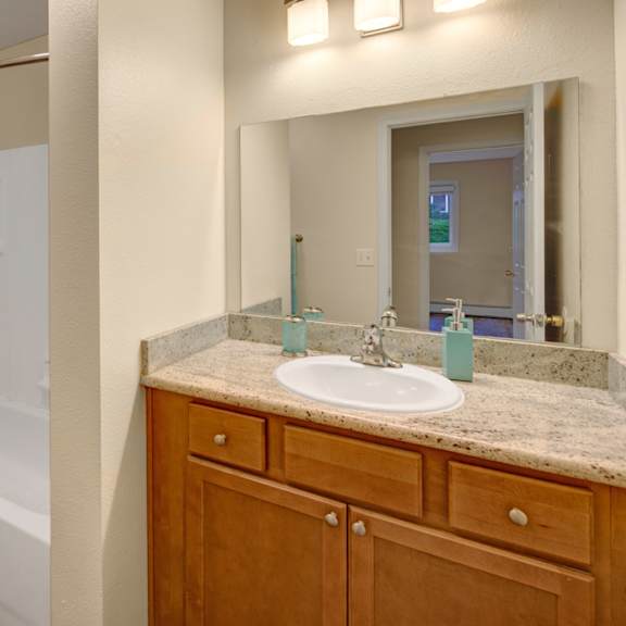 bathroom here at hillside chalet apartments with wooden cabinets and granite countertop, features a sink and large mirror. bathtub with a blue-patterned curtain sits beside a toilet.