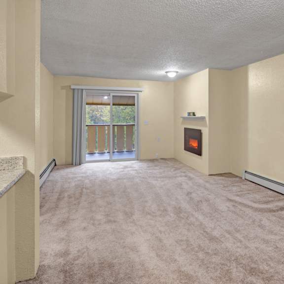 empty living room here at hillside chalet apartments with beige walls and carpet, featuring a corner electric fireplace. sliding glass door leads to a balcony with green foliage outside.