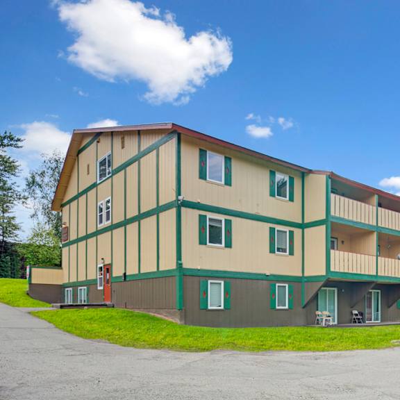 three-story yellow and green hillside chalet apartment building with balconies, set against a bright blue sky. the foreground shows a grassy area and parking lot.
