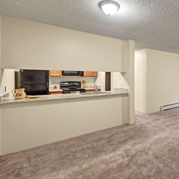 spacious empty living room here at hillside chalet apartments with beige carpet, open kitchen with light wood cabinets, and large sliding glass doors leading to a balcony with greenery.