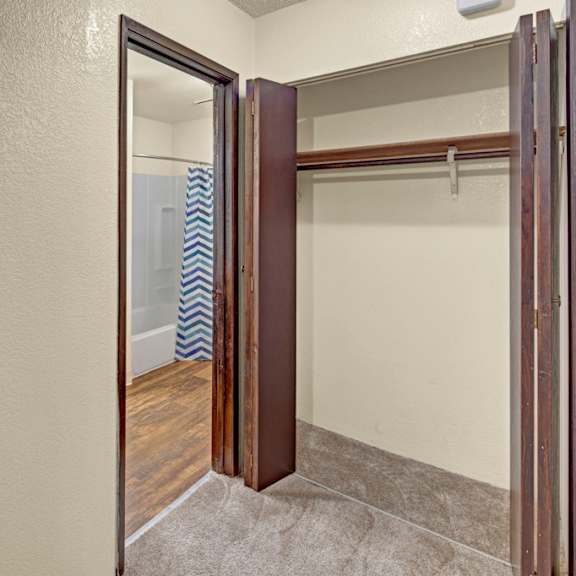 a small, empty walk-in closet here at hillside chalet apartments with beige walls, open wooden shelves, and brown carpet. leads to a bathroom with a chevron-patterned shower curtain.