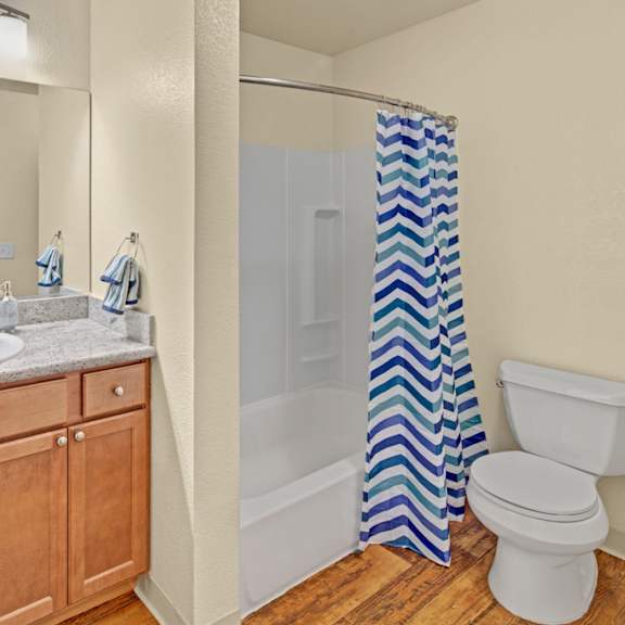 bathroom here at hillside chalet apartments with wooden cabinets, granite countertop, and mirror above a sink. a shower with a blue chevron curtain and a toilet beside it. cozy ambiance.