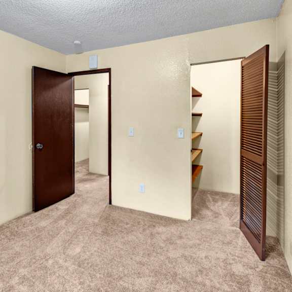 empty beige bedroom here at hillside chalet apartments with brown carpet, featuring an open door leading to another space with shelving. the atmosphere is simple and neutral.
