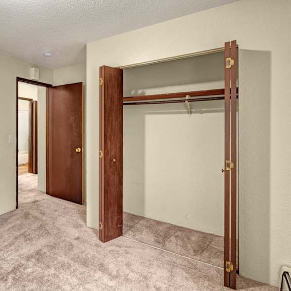 empty bedroom here at hillside chalet apartments with textured beige walls and carpeted floor. an open closet with wooden doors is on the right, and a doorway leads to a bathroom.