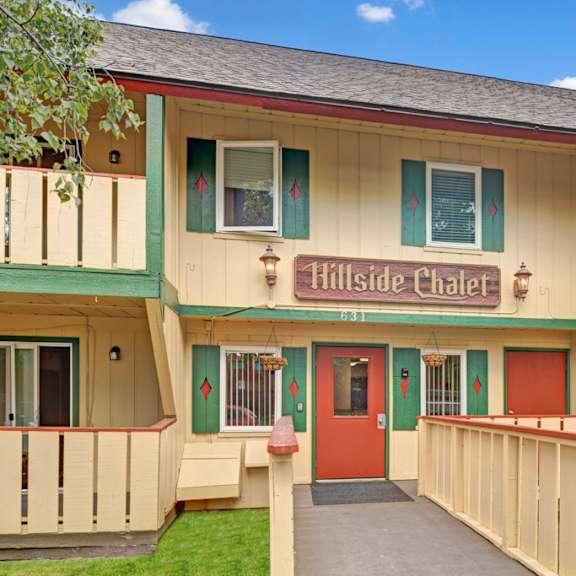 two-story hillside chalet apartment building with a rustic design, labeled "hillside chalet." features balconies with potted plants, a wooden fence, and an american flag.