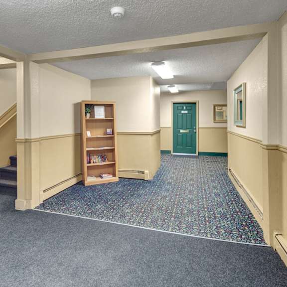 a quiet, well-lit hallway here at hillside chalet apartments with cream-colored walls and patterned carpet. a green door is at the end, with stairs and a bookshelf on the left.