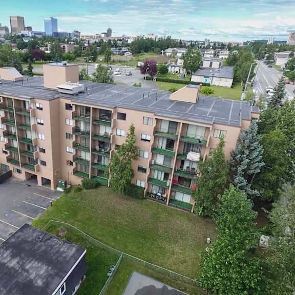 aerial view of a beige, five-story outlook apartment building with balconies, surrounded by green trees and a parking lot. city skyline in the distance under a cloudy blue sky.