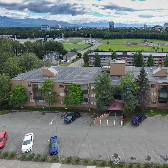 aerial view of a brown outlook apartment building with a parking lot in front, surrounded by trees. in the background, a sports field, trees, and a cloudy sky with distant mountains.