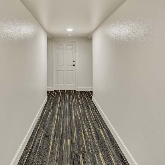 narrow, brightly lit hallway at la maisonnette apartments with white walls and door at the end. the floor features dark patterned carpeting with subtle yellow stripes, creating a modern feel.