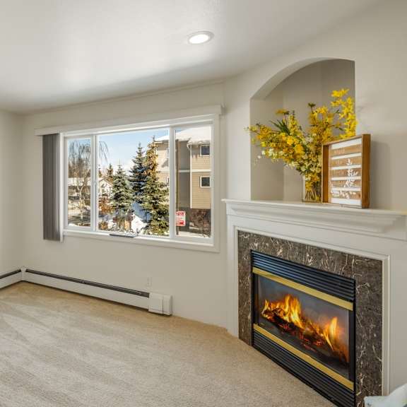 bright living room here at la maisonnette apartments with plush carpet, large window showing snowy outside, and a modern fireplace. yellow flowers and decor on mantel creating a cozy ambiance.