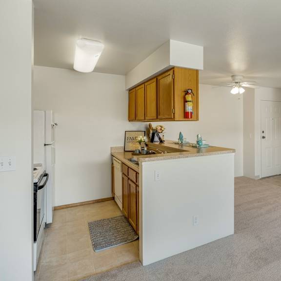 cozy apartment interior here at la maisonette apartments featuring a small kitchen with wooden cabinets, beige countertops, and a dining bar. adjacent is a carpeted living space, with closed white-paneled doors and visible lighting, creating a warm and inviting atmosphere.