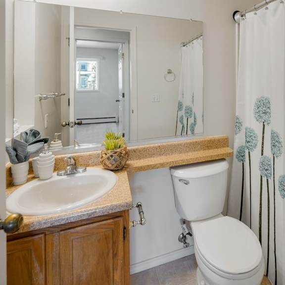 small bathroom here at la maisonnette apartments with wooden vanity and beige countertop. white sink with silver faucet. shower curtain has blue floral design. bright and tidy atmosphere.
