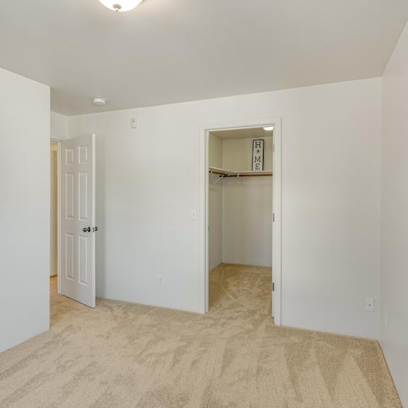 bright bedroom here at la maisonnette apartments with beige carpet and white walls. features an open closet with a "home" sign, and an ajar door leading to another room. calm atmosphere.