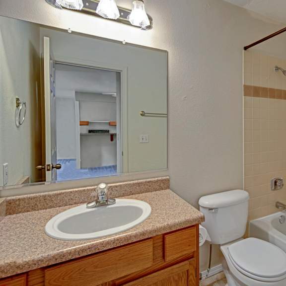 a bathroom here at la maisonnette apartments with beige tiled walls features a bathtub, a white toilet, and a brown vanity with a sink. a mirror reflects a doorway to another room.