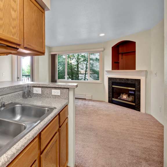 compact kitchen here at la maisonnette apartments with oak cabinets, gray countertops, and stainless steel sink. open view to living room with carpet, large window, and lit fireplace. cozy ambiance.