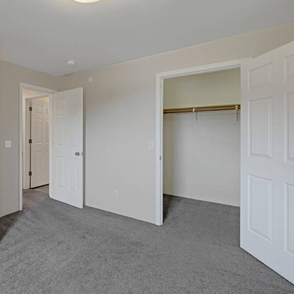 empty bedroom here at la maisonnette apartments with gray carpet and light beige walls. two open doors reveal a closet and a hallway. the space feels clean and minimalistic.