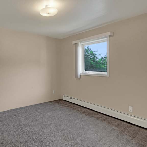 bedroom here at la maisonnette apartments with beige walls, a gray carpet, and a white door ajar. a window with blinds lets natural light in, creating a calm, neutral ambiance.