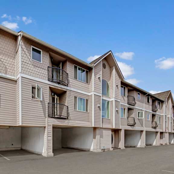 three-story la maisonnette apartment building with beige siding, decorative shingles, and small balconies. open parking space underneath. clear blue sky with a few clouds.